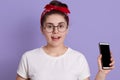 Yong pretty female standing isolated over lilac background looking at camera with excitement, holding phone, wearing casual attire