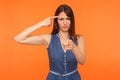 You are idiot. Positive brunette woman in denim dress holding finger near head temple and showing stupid gesture Royalty Free Stock Photo
