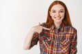 You need me. Smiling redhead woman pointing at herself and looking confident, standing in shirt on white background Royalty Free Stock Photo