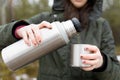 Youg beautifull women pouring beverage from termos to cup in a cold raini day. Healthy living and hiking