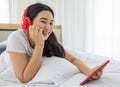 Youn and beautiful asian woman wearing red headphone laying on bed in the bedroom and using tablet computer to work and connect to