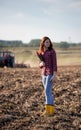 Yound woman holding clipboard monitoring work on plowed field with machinery behind