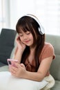 Young adorable Asian girl wearing headphones and using phone, enjoying listening to music on a sofa Royalty Free Stock Photo