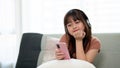 Young adorable Asian girl wearing headphones and using phone, enjoying listening to music on a sofa Royalty Free Stock Photo