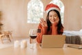 Young adult Asian woman wearing a sweater and santa hat waving in indoor room with glass of wine and candles on table