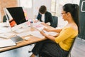 Asian woman using smartphone and desktop computer at modern office, colleague on paperwork in background. People at work concept