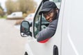 Young adult black delivery man wearing black cap driving white van leaning out the window to look behind. Horizontal Royalty Free Stock Photo