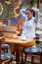 Young adult caucasian female fixing her hair, sitting in the outdoor cafe with a laptop on the table and cup of coffee Royalty Free Stock Photo