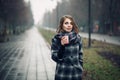 Young adult female with paper cup of coffee staying in city park on cloudy rainy day; Royalty Free Stock Photo