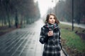 Young adult female with paper cup of coffee staying in city park on cloudy rainy day; Royalty Free Stock Photo