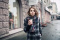 Young adult female with paper cup of coffee staying on the city street on cloudy rainy day; Royalty Free Stock Photo