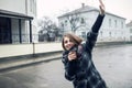 Young adult female with paper cup of coffee staying on the city street on cloudy rainy day and waving; Royalty Free Stock Photo