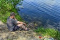 Young adult fisherman sitting on the ground on the shore fishing in a forest pond on a sunny summer-spring day. Man in a gray T- Royalty Free Stock Photo