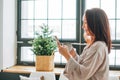 Young adult forty years woman with dark hair in cozy pajamas with cup of tea using mobile phone in living room with Christmas tree