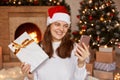 Young adult happy woman wearing white sweater and santa claus hat, sitting near fireplace and xmas tree, expressing positive Royalty Free Stock Photo