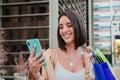 Young adult joyful woman using a cellphone to purchase online on a market place app. Smiling female carrying shopping Royalty Free Stock Photo