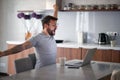 Young adult man with beard and tattoo yawns, stretching in front of laptop on table with cup of coffee and cell phone. lifestyle