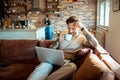 Young adult man using a laptop on a couch in a living room while having a cup of coffee Royalty Free Stock Photo