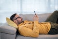 Young adult mature man using smart phone resting lying down on sofa couch at home messaging on mobile phone Guy with eyeglasses Royalty Free Stock Photo