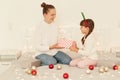 Young adult mother and daughter wearing casual style white sweaters sitting on bed, little girl giving Christmas present to her Royalty Free Stock Photo