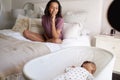 Young adult mother sitting cross legged on her bed looking down at her three month old baby sleeping in his cot, close up Royalty Free Stock Photo