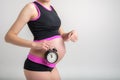 Young adult pregnant girl in sportswear with alarm clock. Time for sports concept. Yoga and fitness in anticipation of childbirth