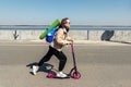 Young adult sporty hipster woman in workout suit, backpack and yoga mat riding kick scooter fast against concrete urban