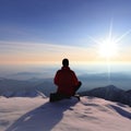 A young adult with a winter jacket is seated in the snow looking out at the majestic mountain range