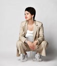 Adult woman in beige business casual pantsuit and sneakers sits squatted looking aside. Stylish business female wear