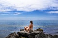 Young adult woman in bikini sits on a granite stone and looks at the sea - view from the back. European female tourist sits on a