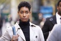 Young adult woman wearing turtleneck sweater and mackintosh walking on a busy street in London, front view