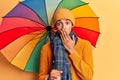 Young african amercian man holding colorful umbrella covering mouth with hand, shocked and afraid for mistake