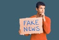 Young african amercian man holding fake news banner serious face thinking about question with hand on chin, thoughtful about