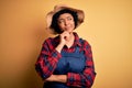 Young African American afro farmer woman with curly hair wearing apron and hat with hand on chin thinking about question, pensive