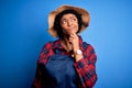 Young African American afro farmer woman with curly hair wearing apron and hat with hand on chin thinking about question, pensive