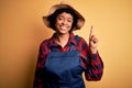 Young African American afro farmer woman with curly hair wearing apron and hat showing and pointing up with finger number one Royalty Free Stock Photo