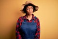 Young African American afro farmer woman with curly hair wearing apron and hat smiling looking to the side and staring away Royalty Free Stock Photo