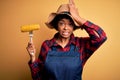 Young African American afro farmer woman with curly hair wearing apron holding cob corn stressed with hand on head, shocked with Royalty Free Stock Photo