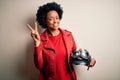 Young African American afro motorcyclist woman with curly hair holding motorcycle helmet smiling with happy face winking at the