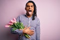 Young african american afro romantic man with dreadlocks holding bouquet of pink tulips sticking tongue out happy with funny Royalty Free Stock Photo