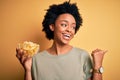 Young African American afro woman with curly hair holding bowl with chips potatoes pointing and showing with thumb up to the side