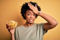 Young African American afro woman with curly hair holding bowl with chips potatoes stressed with hand on head, shocked with shame Royalty Free Stock Photo