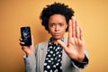 Young African American afro woman with curly hair holding cracked broken smartphone with open hand doing stop sign with serious