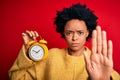 Young African American afro woman with curly hair holding vintage alarm clock with open hand doing stop sign with serious and Royalty Free Stock Photo