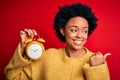 Young African American afro woman with curly hair holding vintage alarm clock pointing and showing with thumb up to the side with Royalty Free Stock Photo