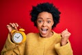 Young African American afro woman with curly hair holding vintage alarm clock screaming proud and celebrating victory and success Royalty Free Stock Photo