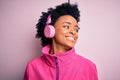 Young African American afro woman with curly hair listening to music using pink headphones looking away to side with smile on Royalty Free Stock Photo