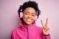 Young African American afro woman with curly hair listening to music using pink headphones smiling with happy face winking at the Royalty Free Stock Photo