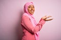 Young African American afro woman wearing muslim hijab over isolated pink background pointing aside with hands open palms showing Royalty Free Stock Photo