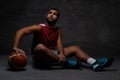 Young African-American basketball player in sportswear sitting on a floor with a ball on a dark background. Royalty Free Stock Photo